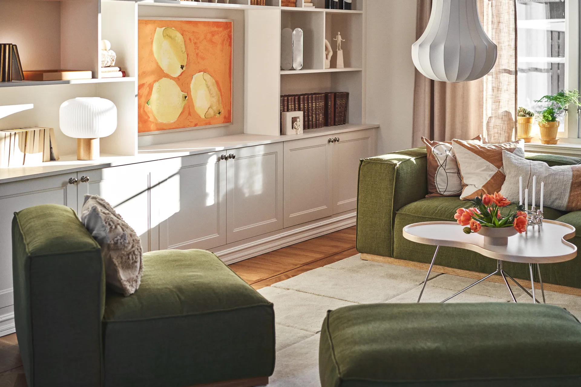 Solrikt stue med grønne sofaer, et hvitt salongbord med oransje tulipaner og lys, samt hvite innebygde hyller med kunst.