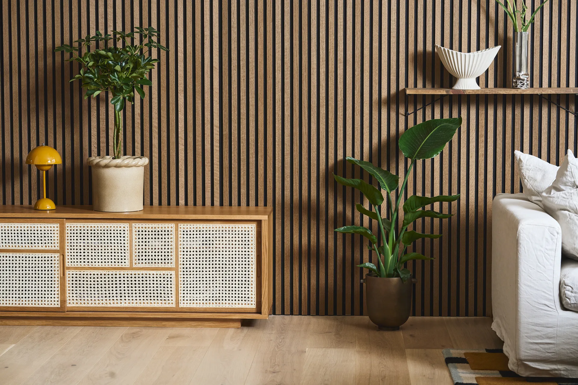 Moderne stue med spilevegg i tre, rottingkommode, gul lampe, potteplanter og en hvit sofa.