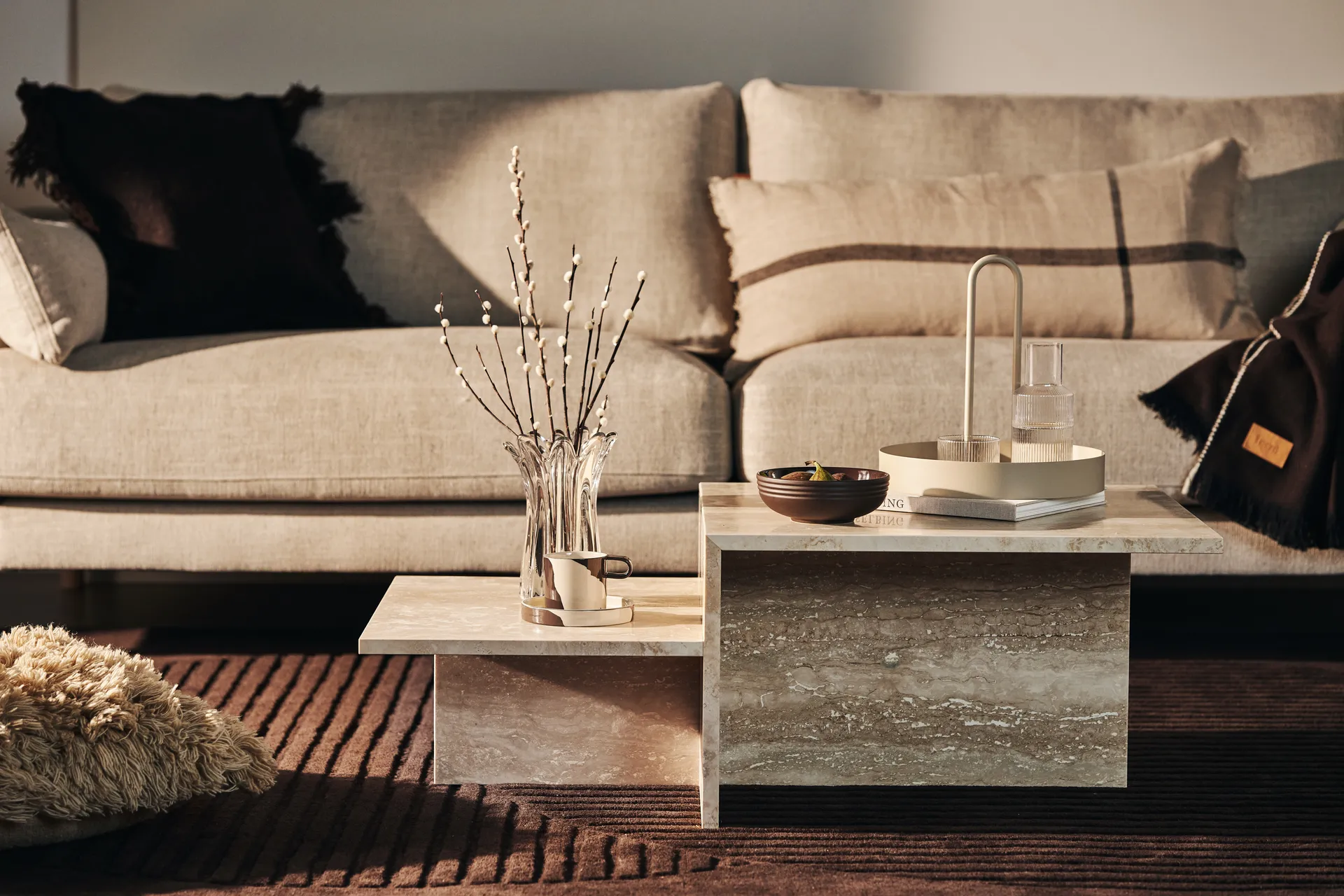 Nivådelt sofabord i beige stein foran en nøytral sofa med dekorativt tilbehør.