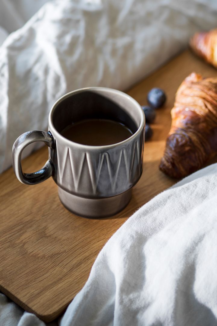 Espressokoppen Coffee & More i grått står på et trefat i en seng, med en croissant ved siden av.
