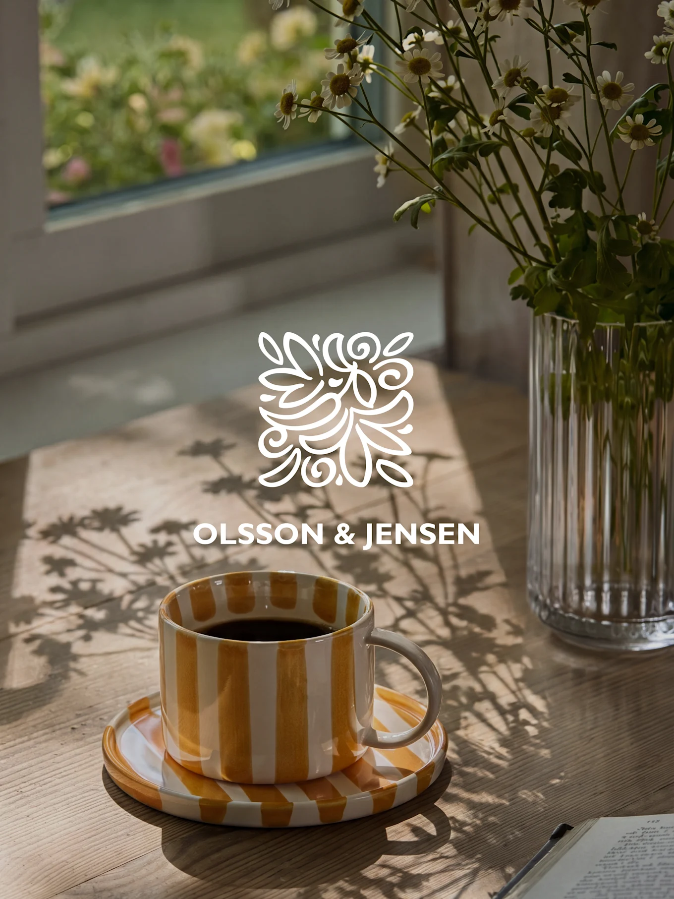 Gul- og hvitstripet kaffekopp på et trebord med en vase med hvite blomster og solbelyste skygger, Olsson & Jensen-logo.