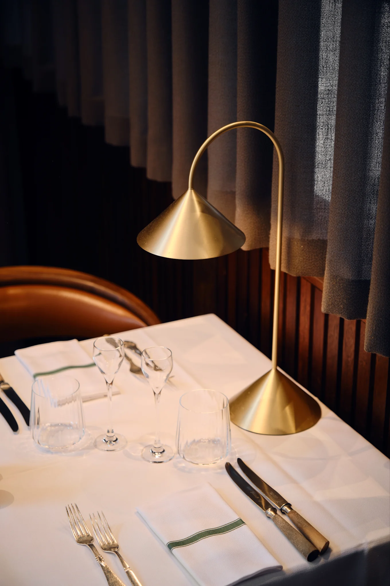 En moderne gullbordlampe lyser opp et dekket restaurantbord med hvite duker, glass, bestikk og en serviett med grønn stripe.