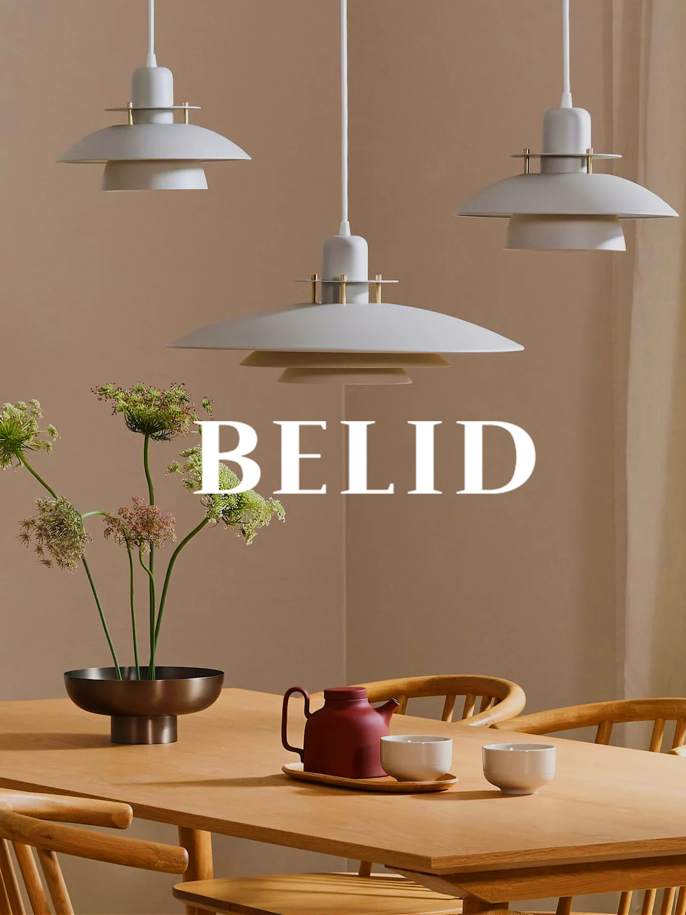 Tre lagdelte lysegrå pendellamper over et spisebord i tre med blomster, en rød tekanne og kopper. Merkenavn BELID.