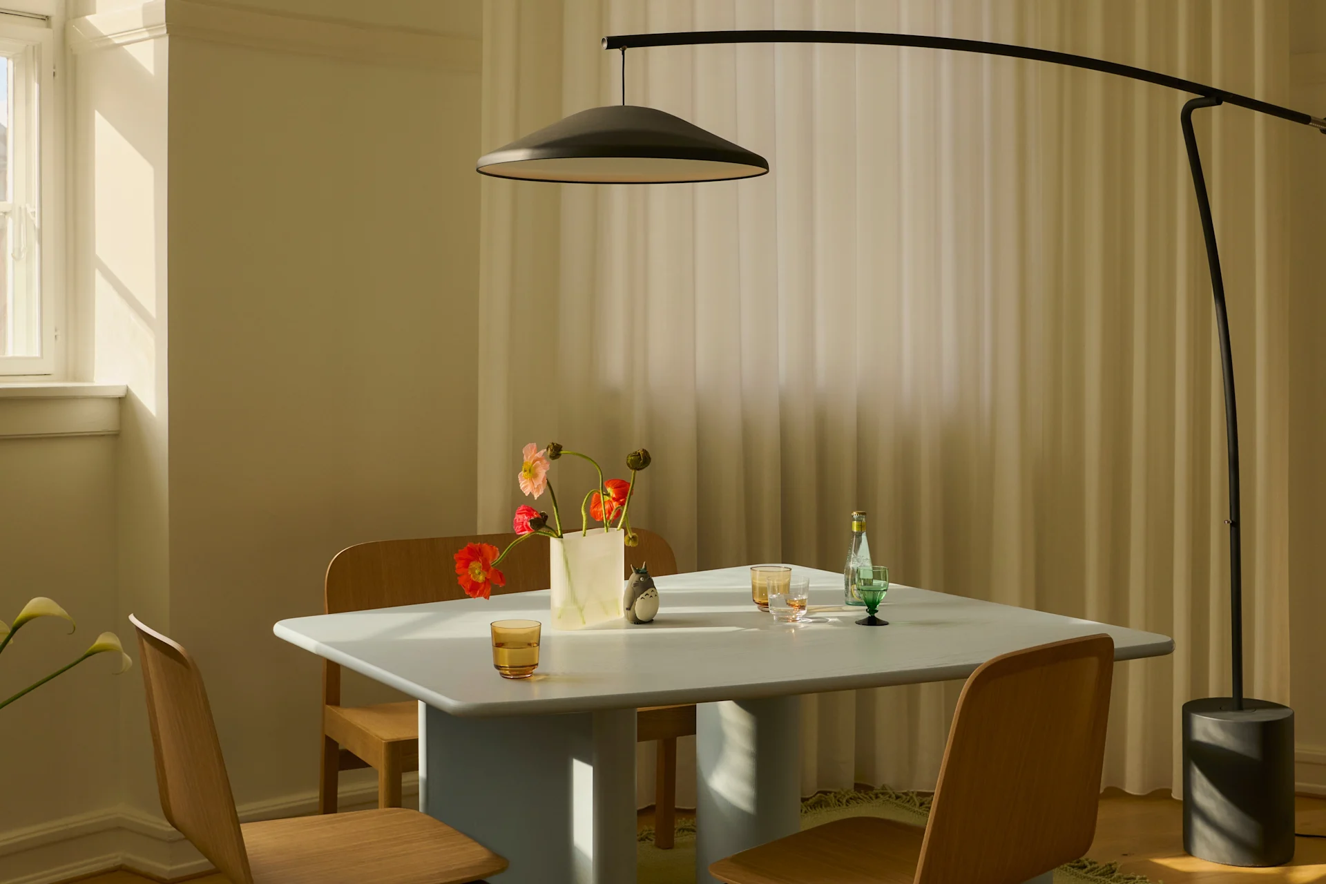 Et moderne spisestue med et lyseblått bord, trestoler, en svart buet gulvlampe og en vase med fargerike blomster i sollys.