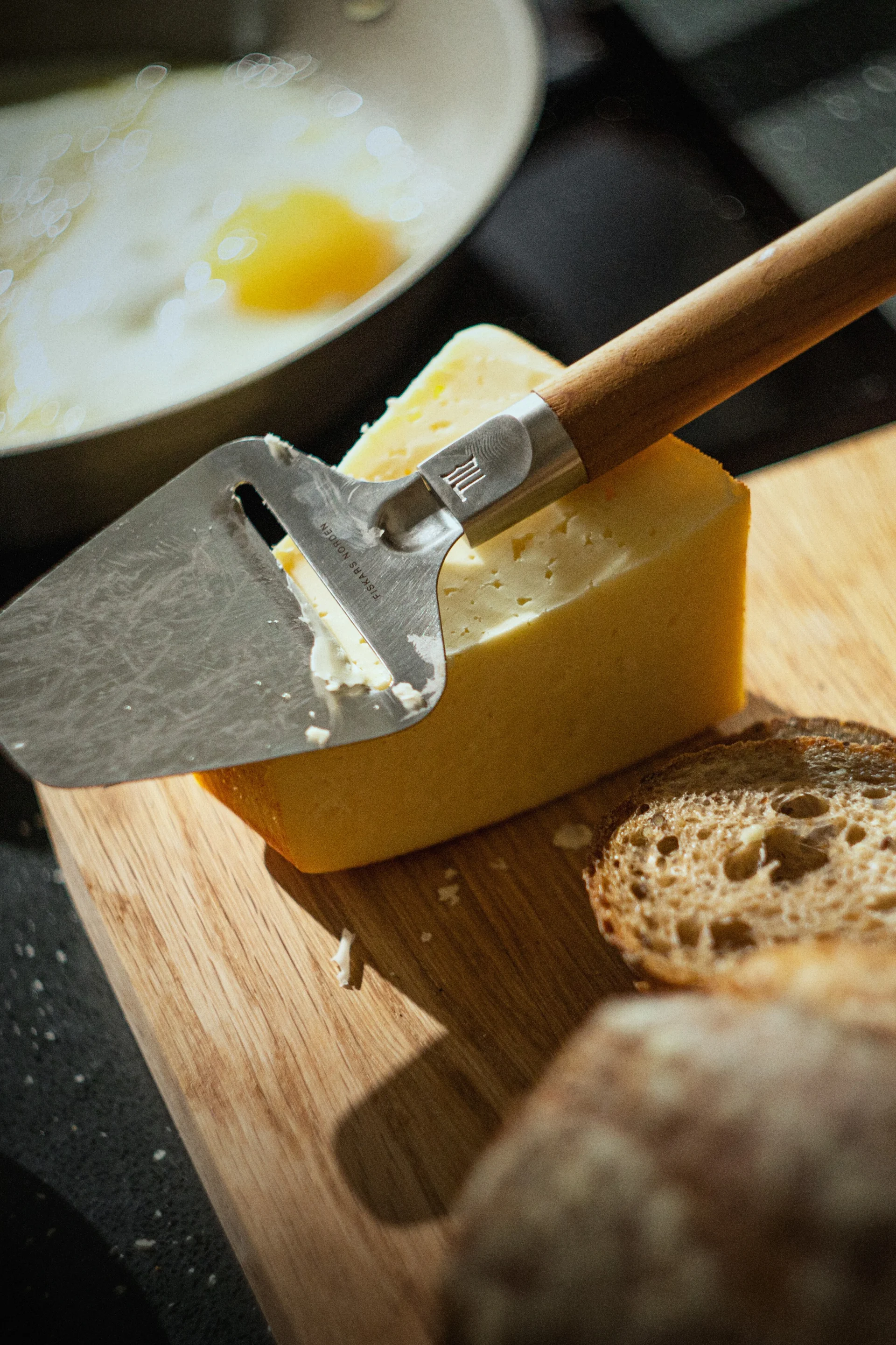 En osteskjærer hviler på en blokk gul ost på et skjærebrett av tre, med brødskiver og et stekt egg i en panne i bakgrunnen.