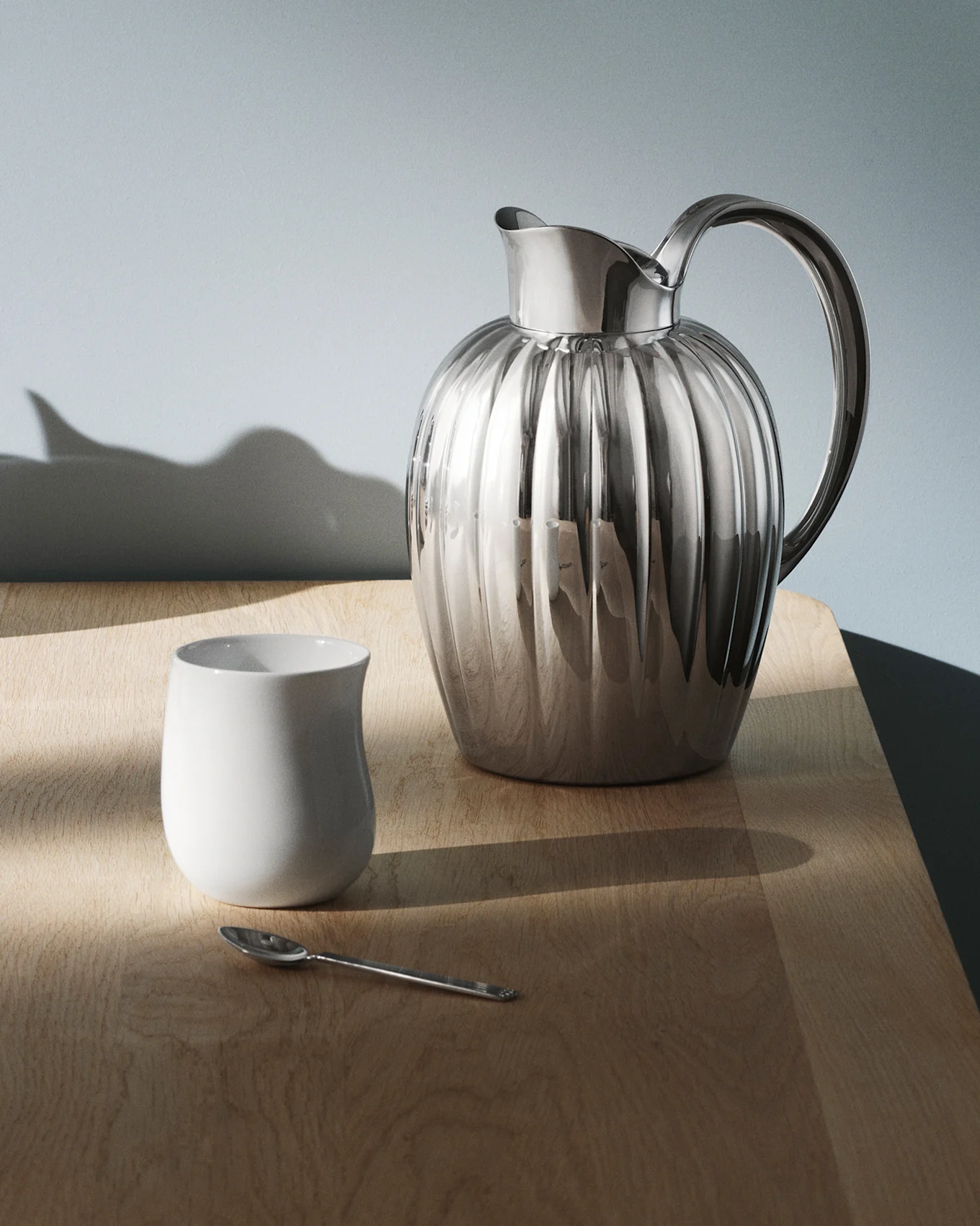 En skinnende, riflet sølvkanne med hank står på et mørkt, reflekterende bord, med lysmønstre kastet over overflaten.