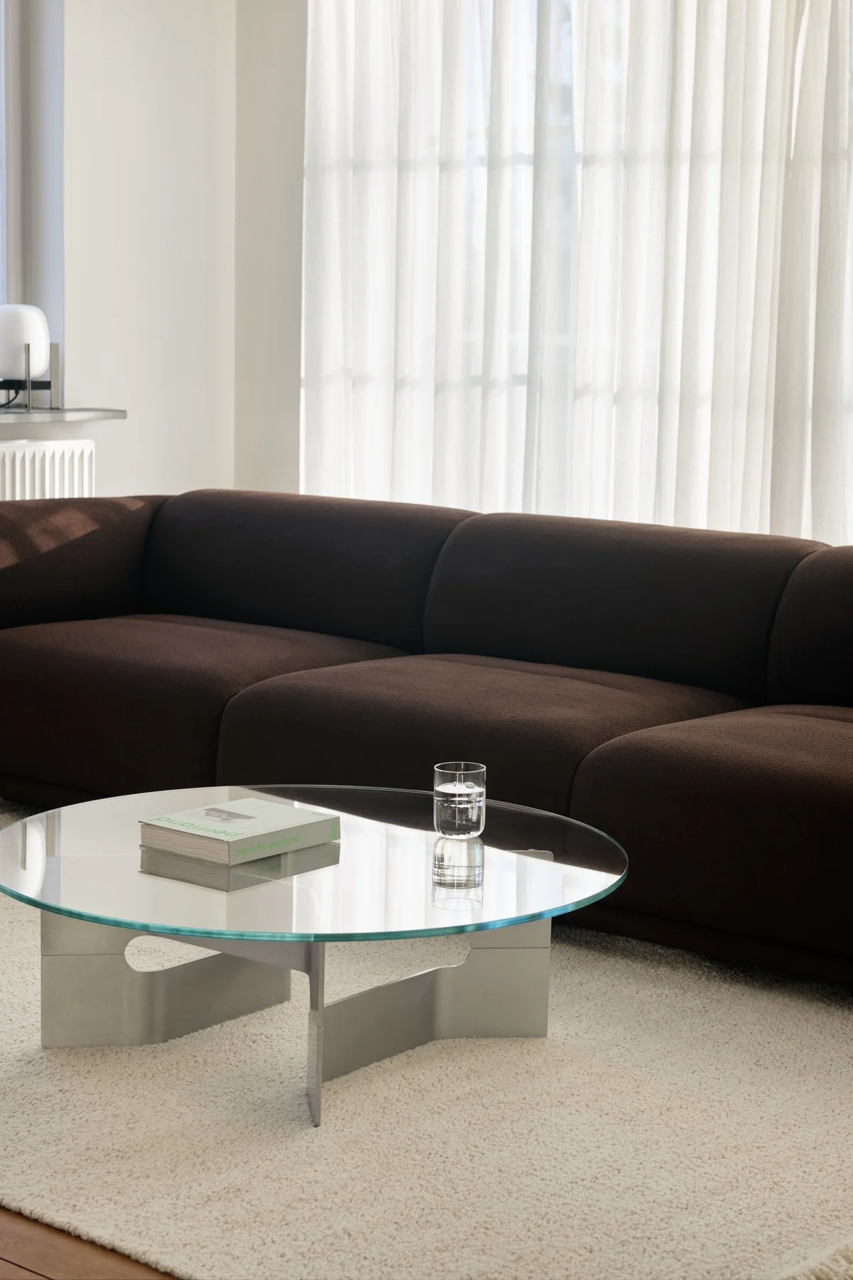 Moderne stue med mørkebrun modulsofa, rundt glass sofabord og lyst, raggete teppe.