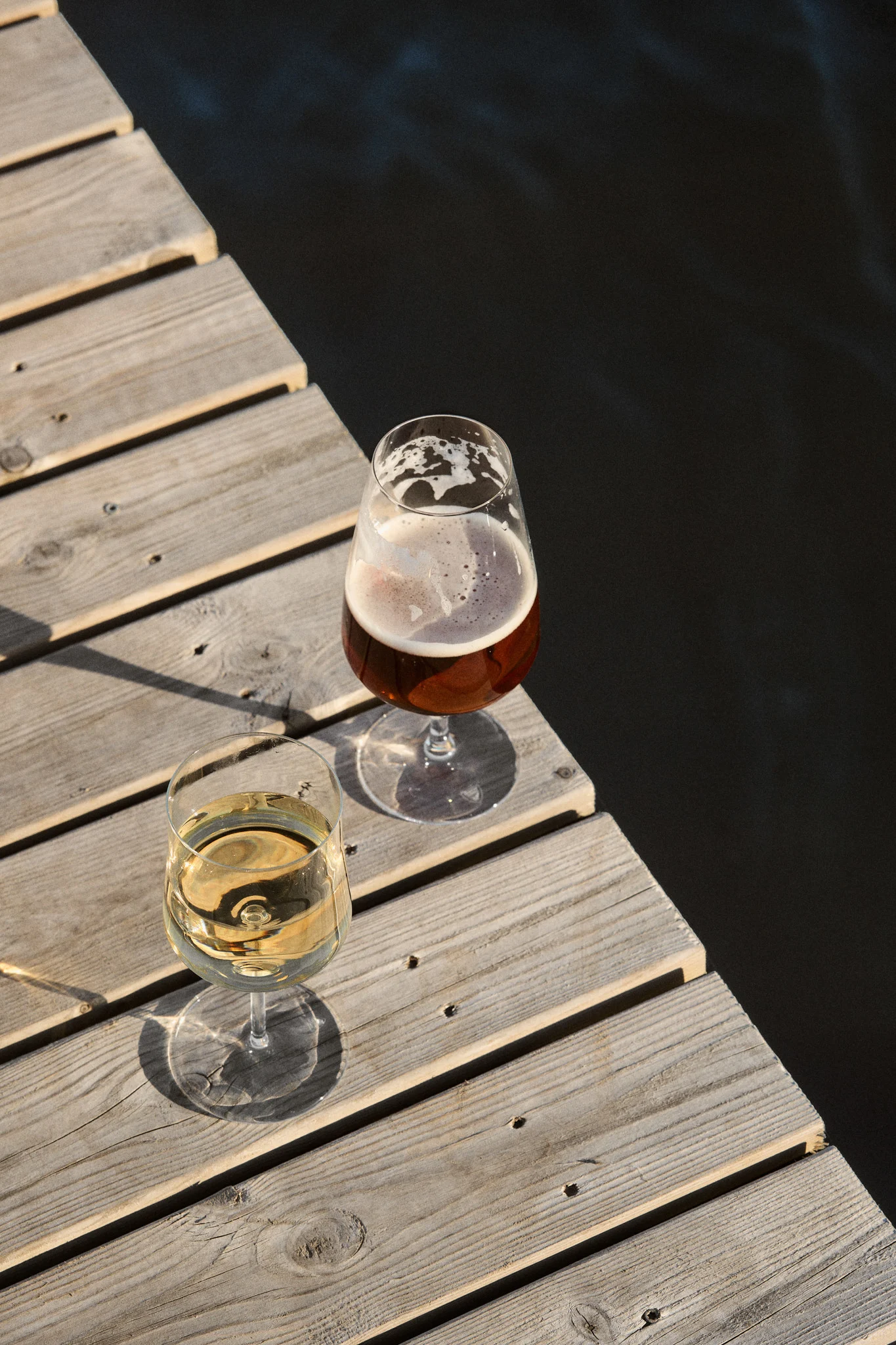 To glass, et med hvitvin og et med øl, på en trebrygge ved mørkt vann i sollys.