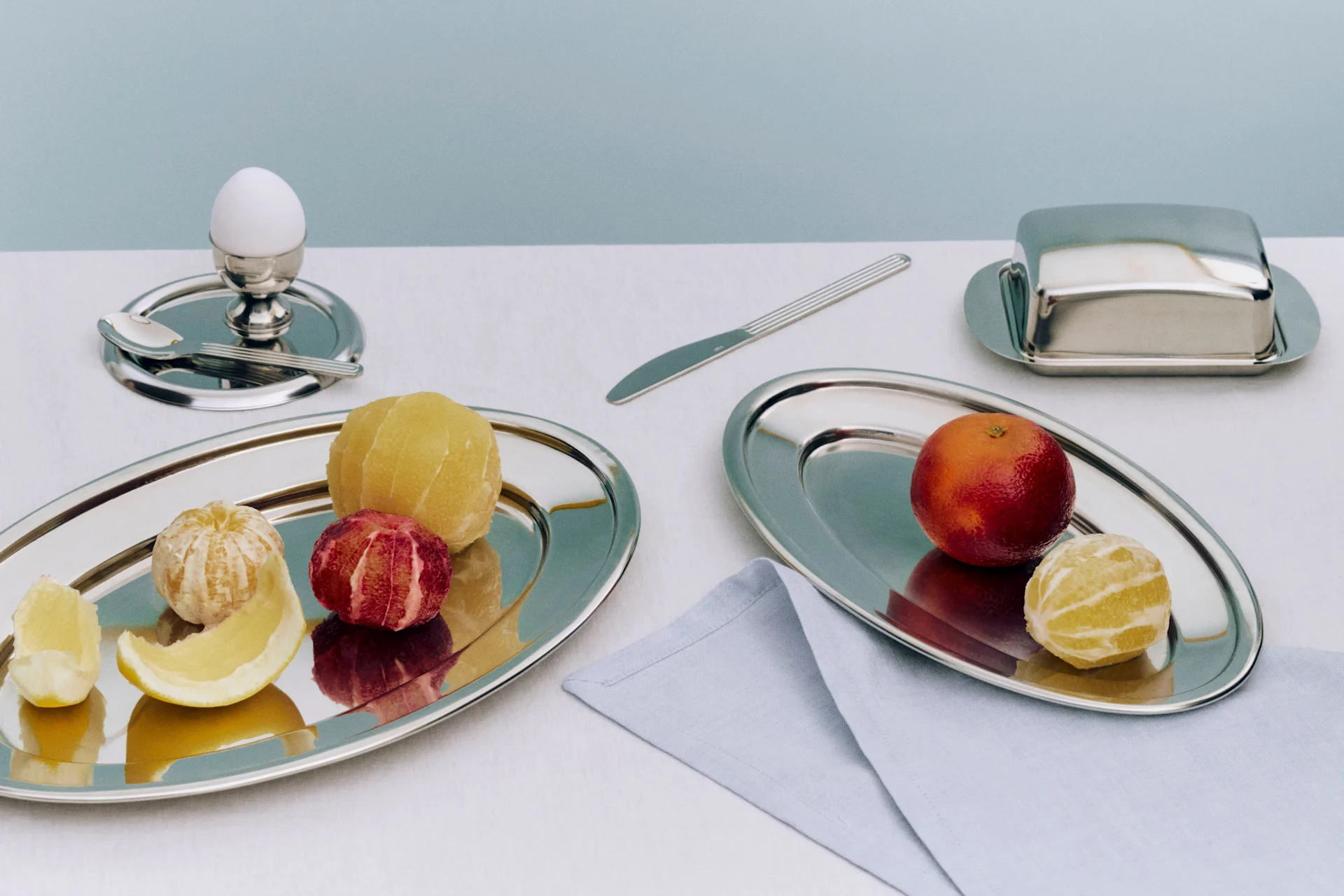 Frokost med skrellede og uskrellede sitrusfrukter på sølvfat, et egg i eggeglass og en smørboks.