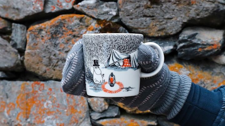 @moominbyingri holder Sleep Well-mummikoppen foran en steinmur, iført en hanske.
