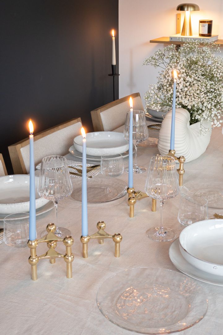 Lyseblå antikke lys fra Ester & Erik er plassert i gyldne lysestaker fra STOFF på et middagsbord med hvitt servise og en beige duk.