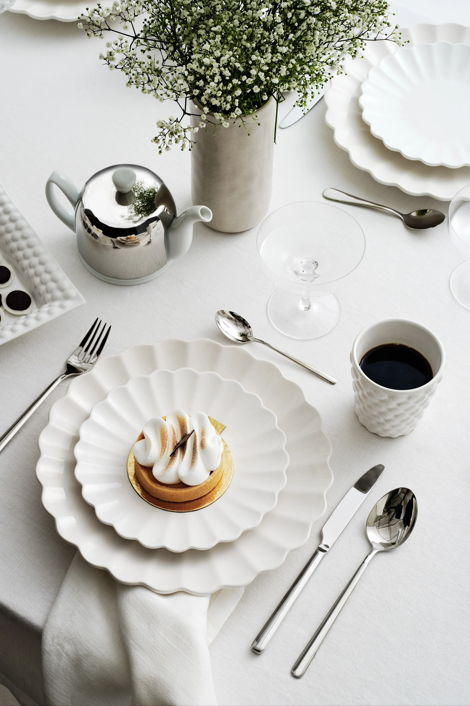 Elegant borddekking med hvite bølgede tallerkener, en marengsterte, kaffe, en sølv tekanne og brudeslørblomster.