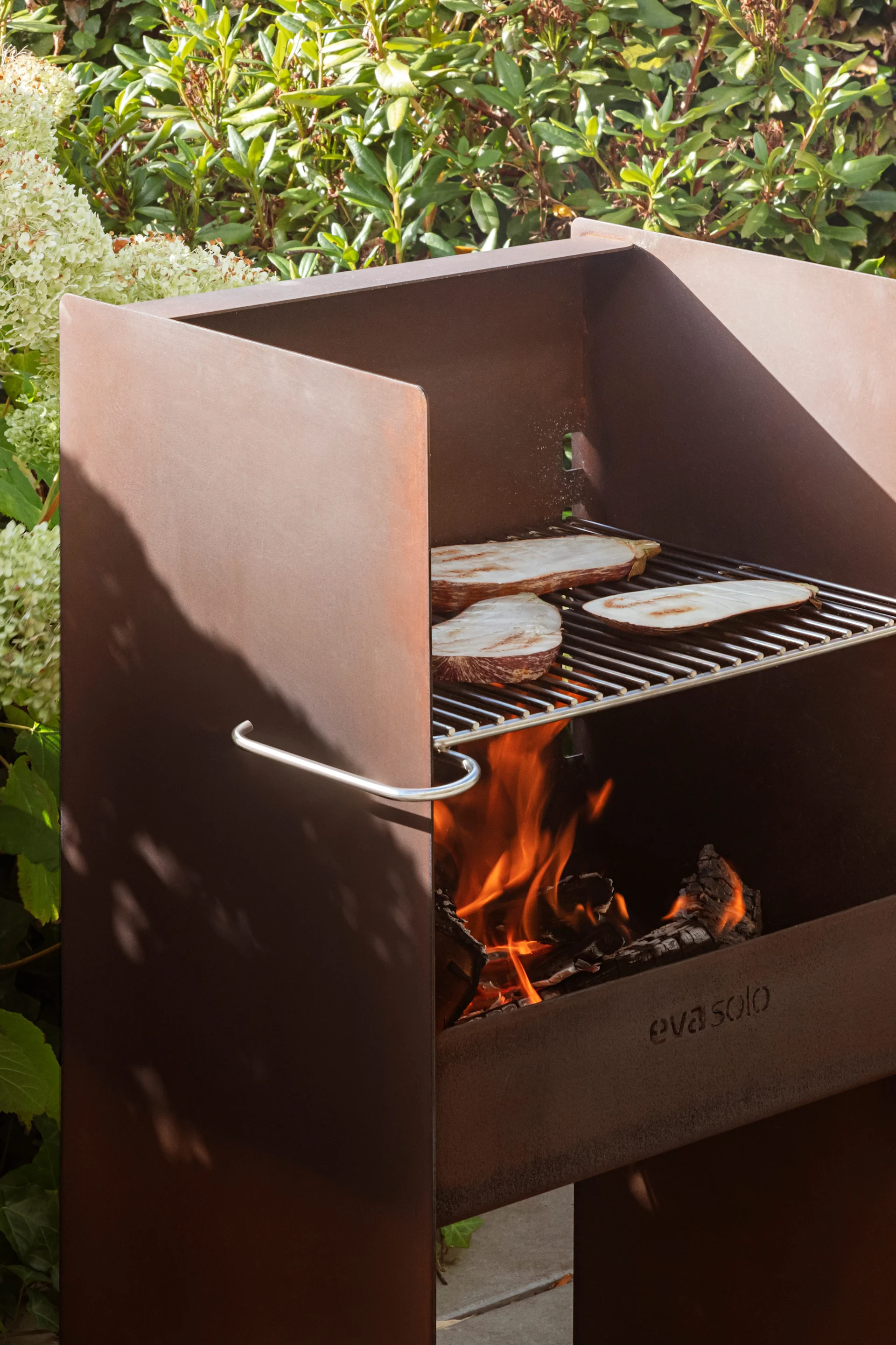 Utendørs grill i cortenstål med aubergineskiver som grilles på rist over åpen ild, omgitt av grønne planter.