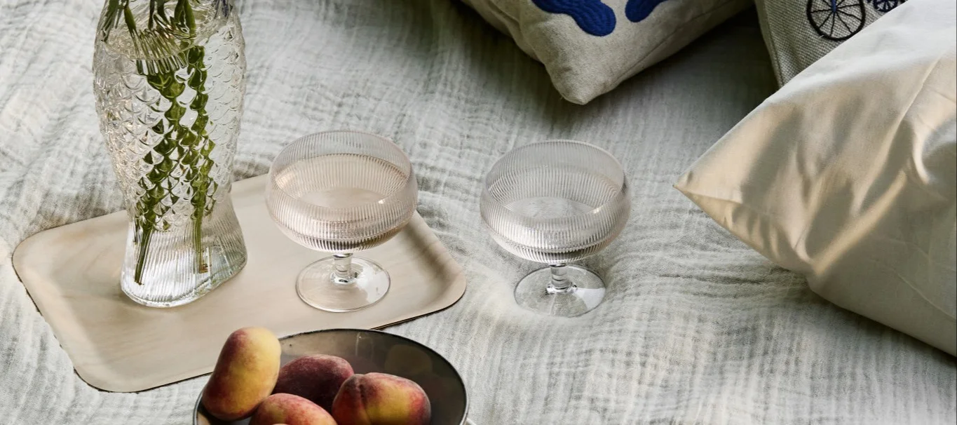 Piknikoppsett med fiskeformet vase, to riflede glass og fersken på et lyst teppe.