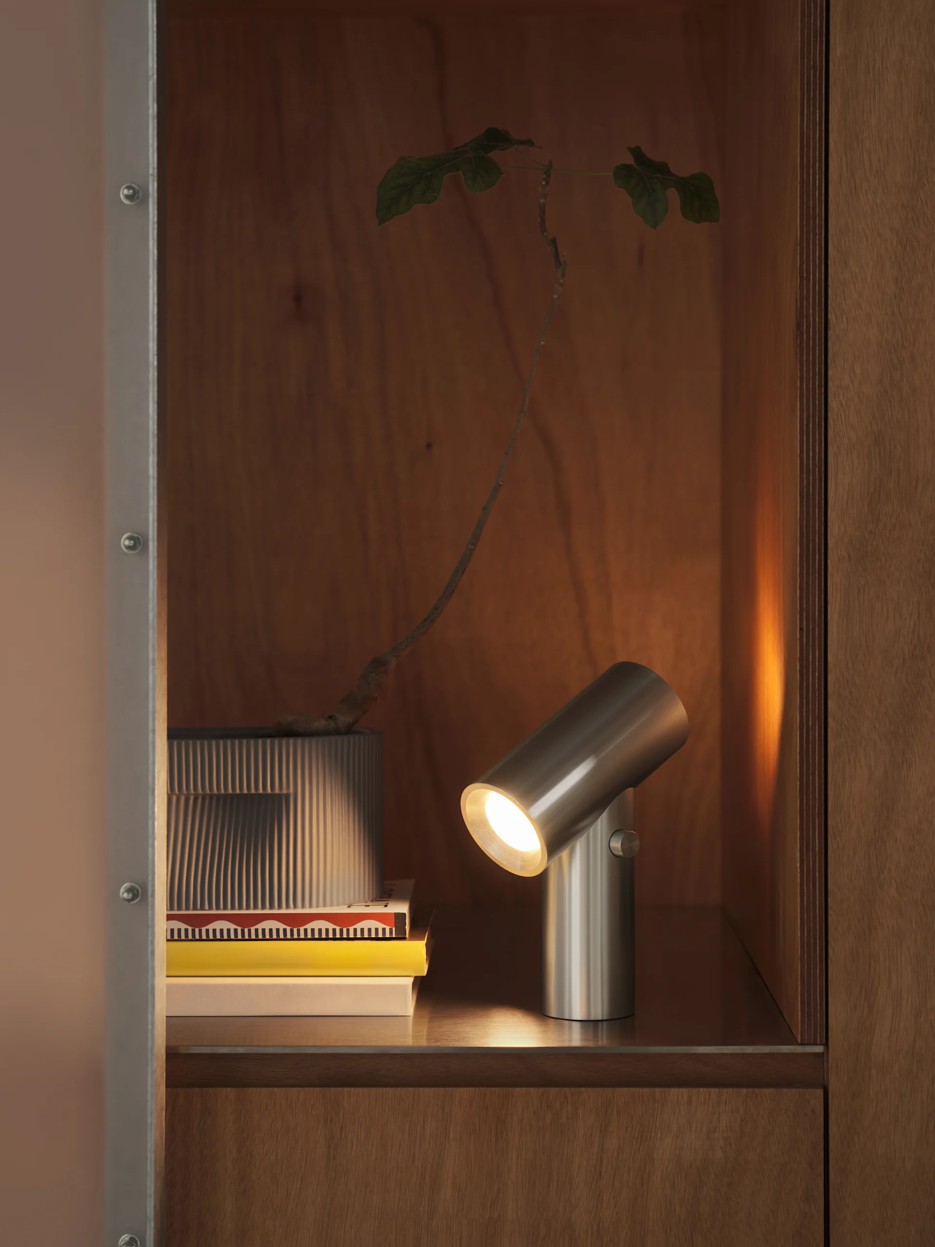 En sylindrisk lampe i børstet metall lyser opp en stabel bøker og en plante i en riflet potte, inne i et treskap.