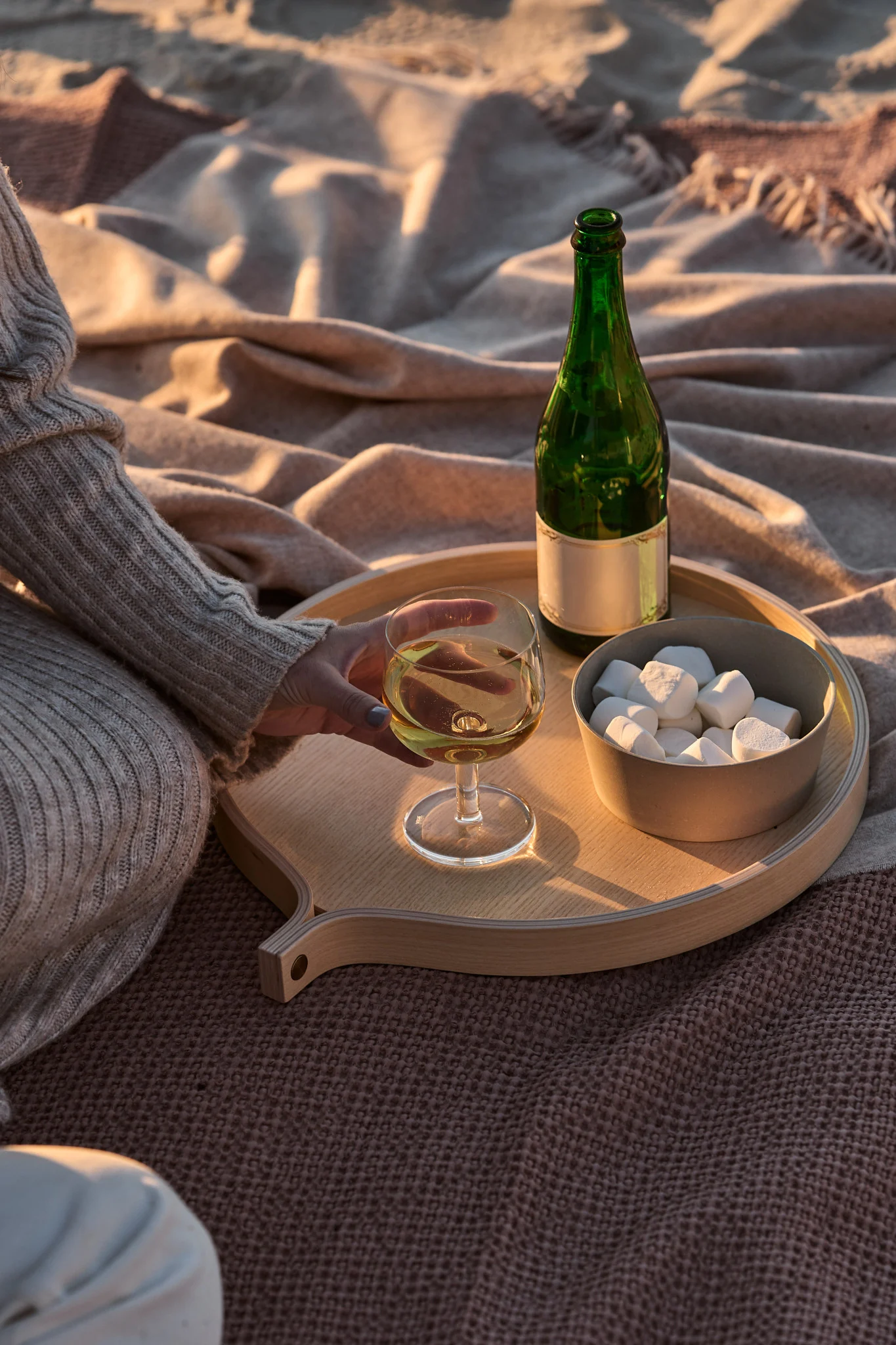 Person holder hvitvin på et trebrett med grønn flaske og marshmallows på tepper i den gyldne time.