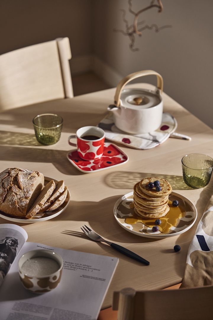 Et frokostbord dekket med Marimekko Unikko tallerkener og tekanne og en stabel pannekaker med sirup.