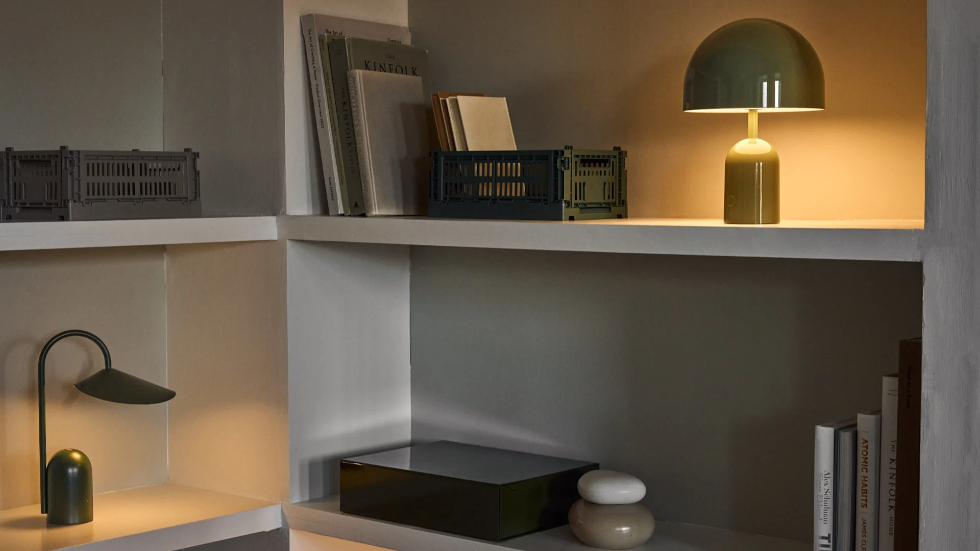 Hjørne med hyller, en grønn kuppelformet bordlampe, en grønn buet bordlampe, sammenleggbare kasser og bøker.