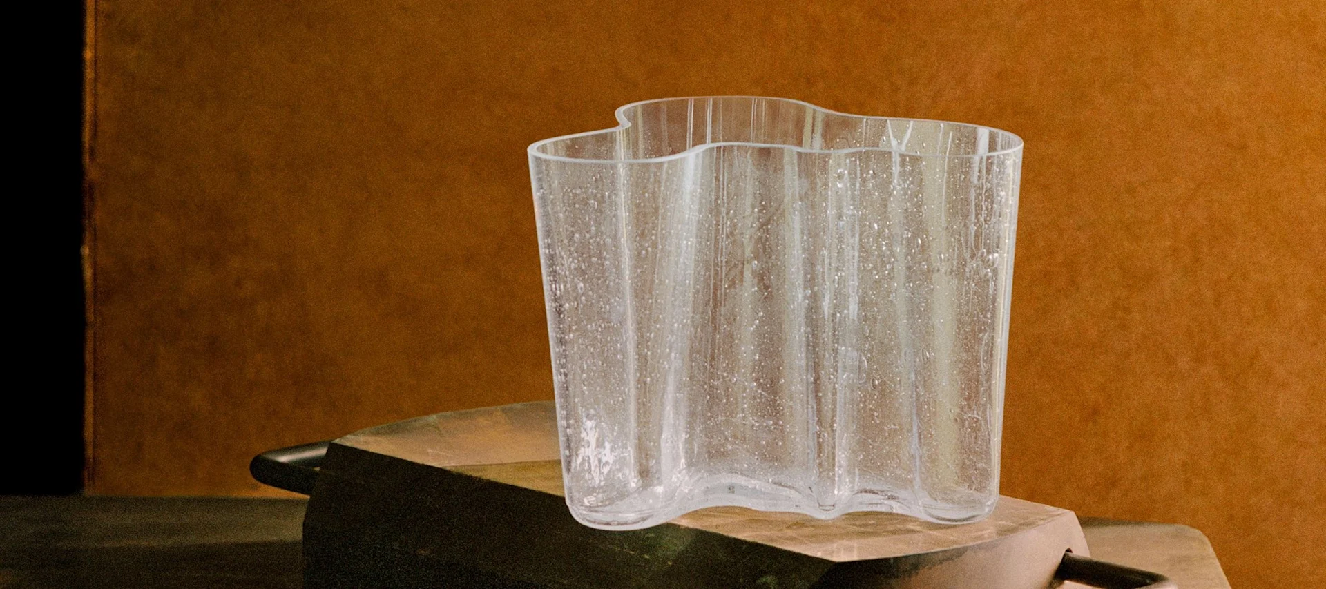 En klar, bølget glassvase med teksturerte flekker på en rustikk mørk overflate mot en varm brun bakgrunn.