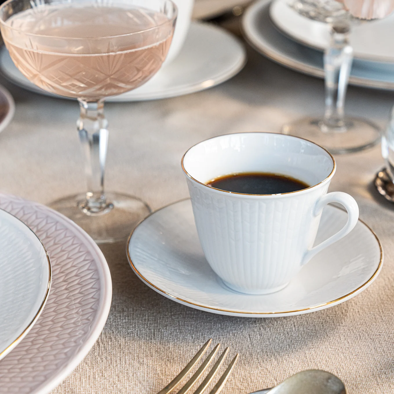 En hvit kaffekopp med gullkant på en skål, ved siden av en rosa tallerken og et rosévinglass på en linduk.