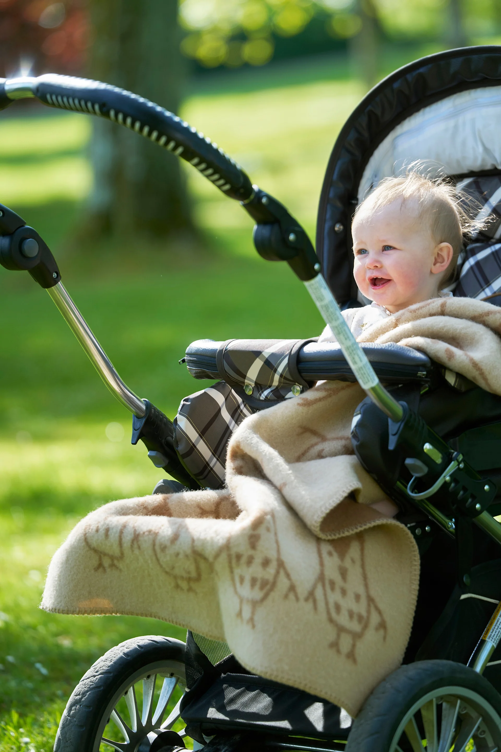 En smilende baby med lyst hår, innpakket i et ugle-mønstret teppe, sitter i en barnevogn utendørs i en solrik park.