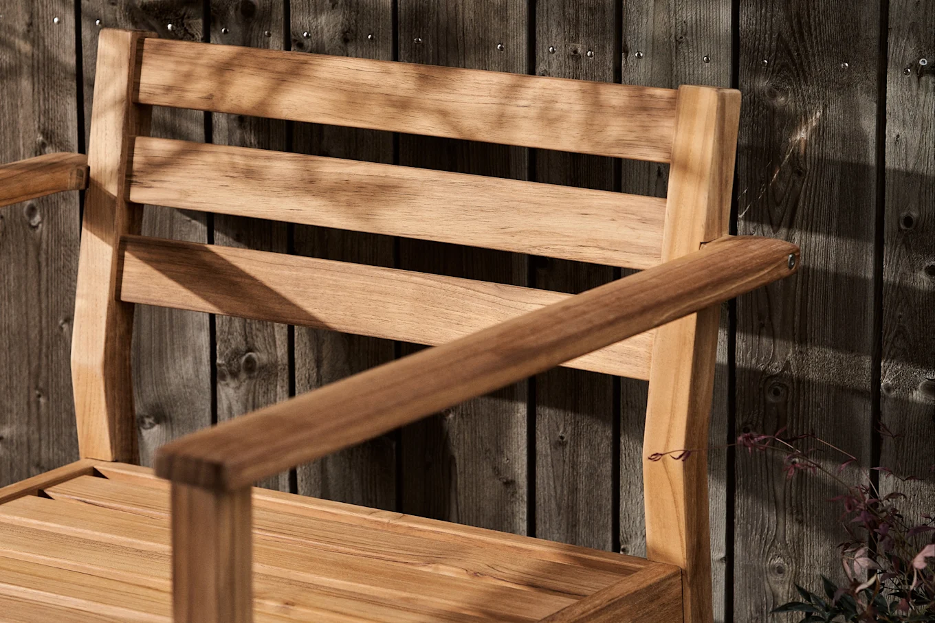 Lamellert trestol med armlener mot et mørkt tregjerde, dekket av bladskygger.