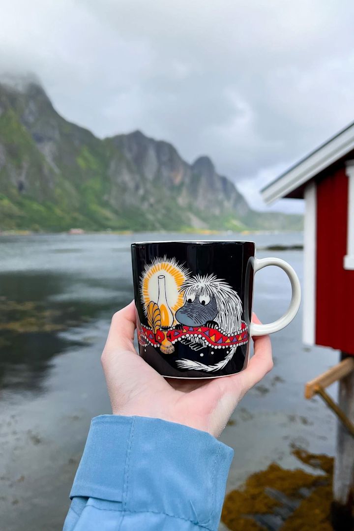 @moominbyingri holder Urahn-mummikoppen i hånden, med et røft norsk landskap i bakgrunnen.
