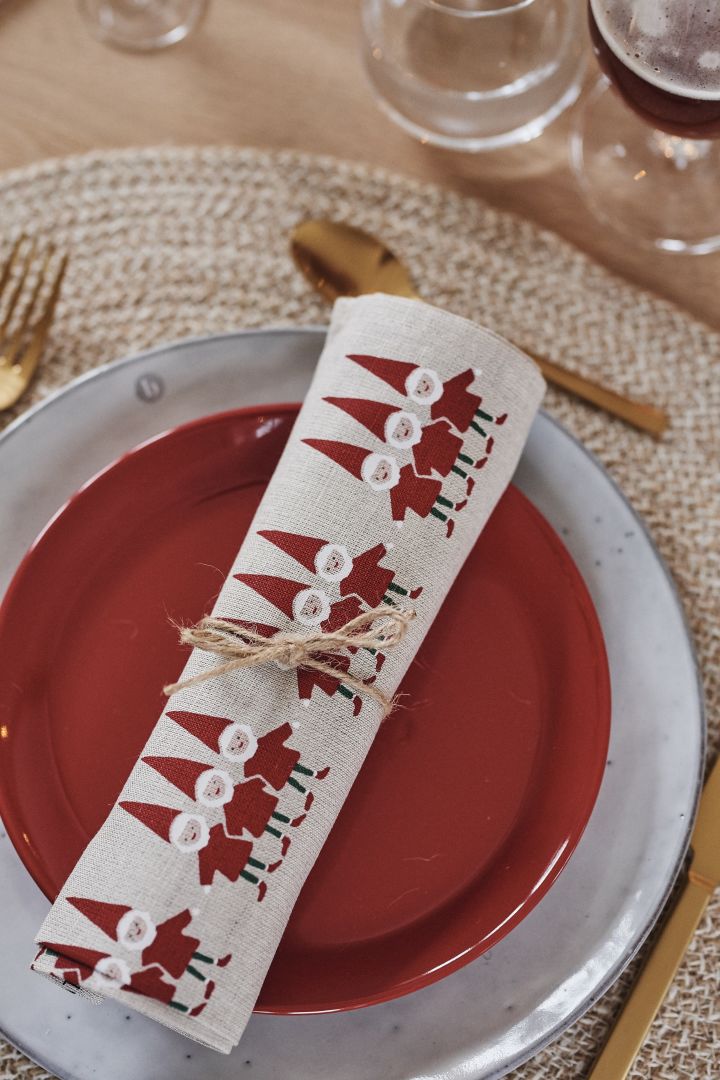 Sammenrullet kjøkkenhåndkle med nissemotiv hviler på en rød asjett og Nordic Sand tallerkenen, som blir kuvert for en rød borddekking til jul.
