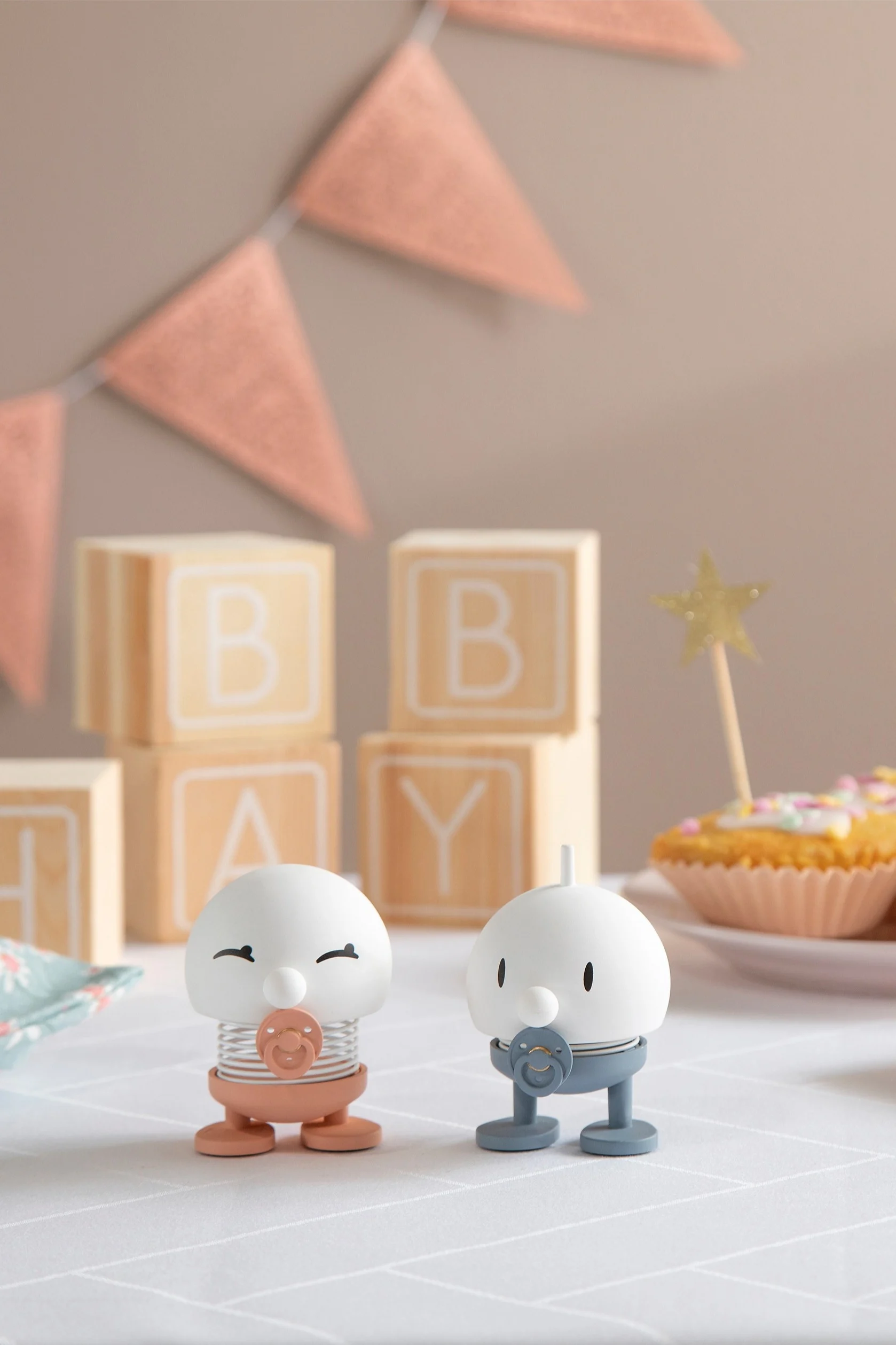 Hoptimist Baby Bimble finnes i rosa og blått og står på et bord pyntet med gaver, kranser og cupcakes.