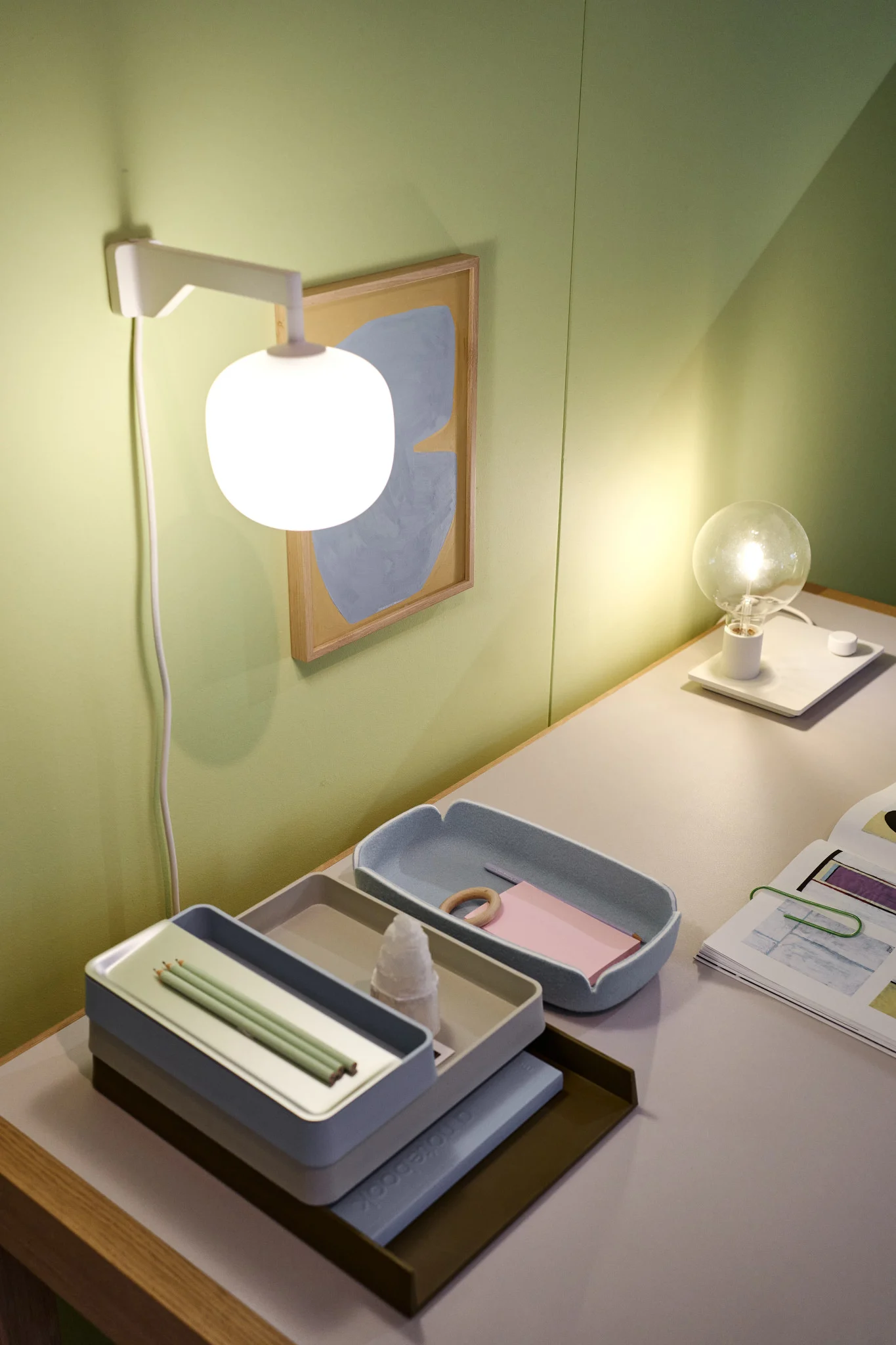 Et ryddig skrivebord foran en grønn vegg, med den beige Rime-vegglampen fra Muuto.