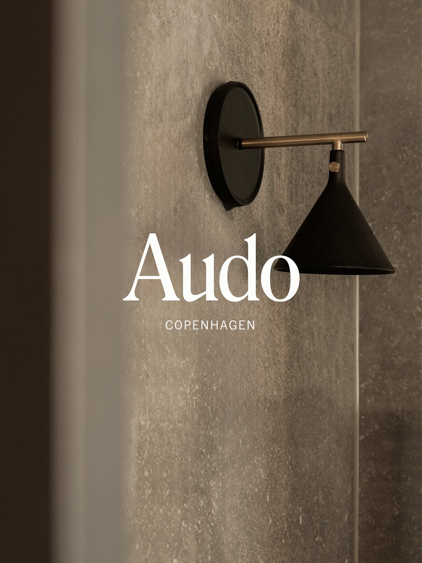 En veggmontert svart lampe med messingarm mot en teksturert grå vegg, med teksten 'Audo Copenhagen'.