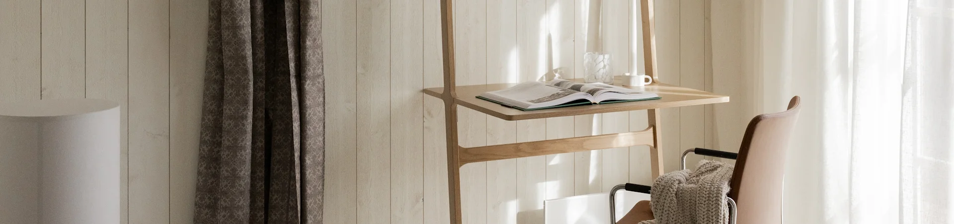 Et trestativ med åpen bok, glassvase og hvitt krus, ved siden av en brun skinnstol med et beige teppe, badet i sollys.