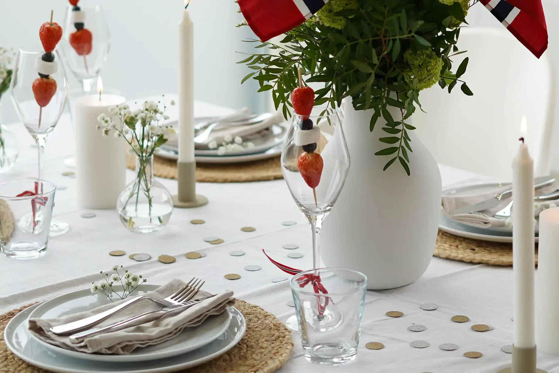 Festbord dekket med hvite tallerkener, lys, blomsteroppsatser, fruktspyd i glass og norske flagg.