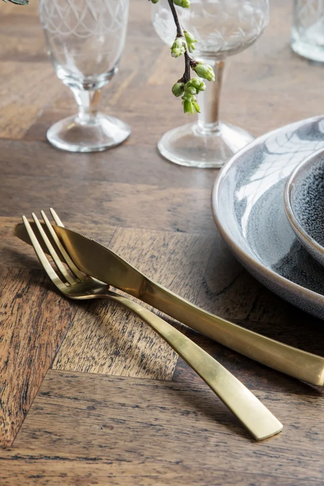 Gullfarget gaffel og kniv på et trebord ved siden av keramiske tallerkener og mønstrede glass, med en liten gren med grønne knopper.