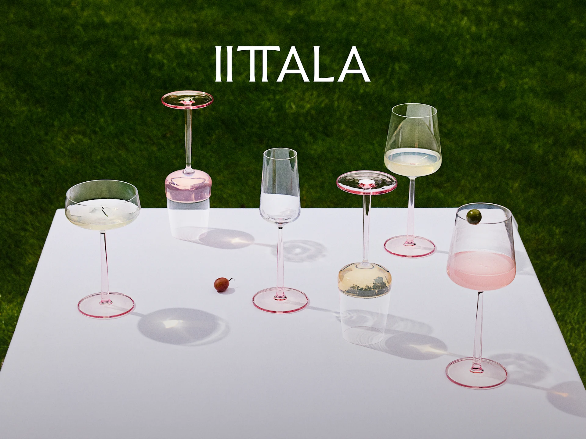 Iittala-logo over en samling vinglass med rosa stilk på et hvitt bord med grønt gress i bakgrunnen, noen med drinker.