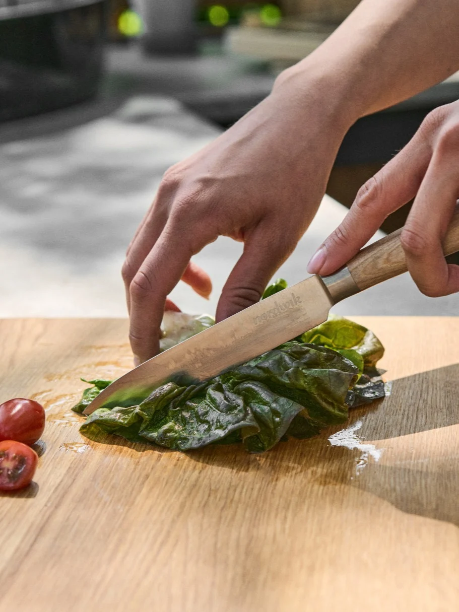 Hender kutter frisk grønn salat på et skjærebrett i tre med en kniv, ved siden av skivede cherrytomater.