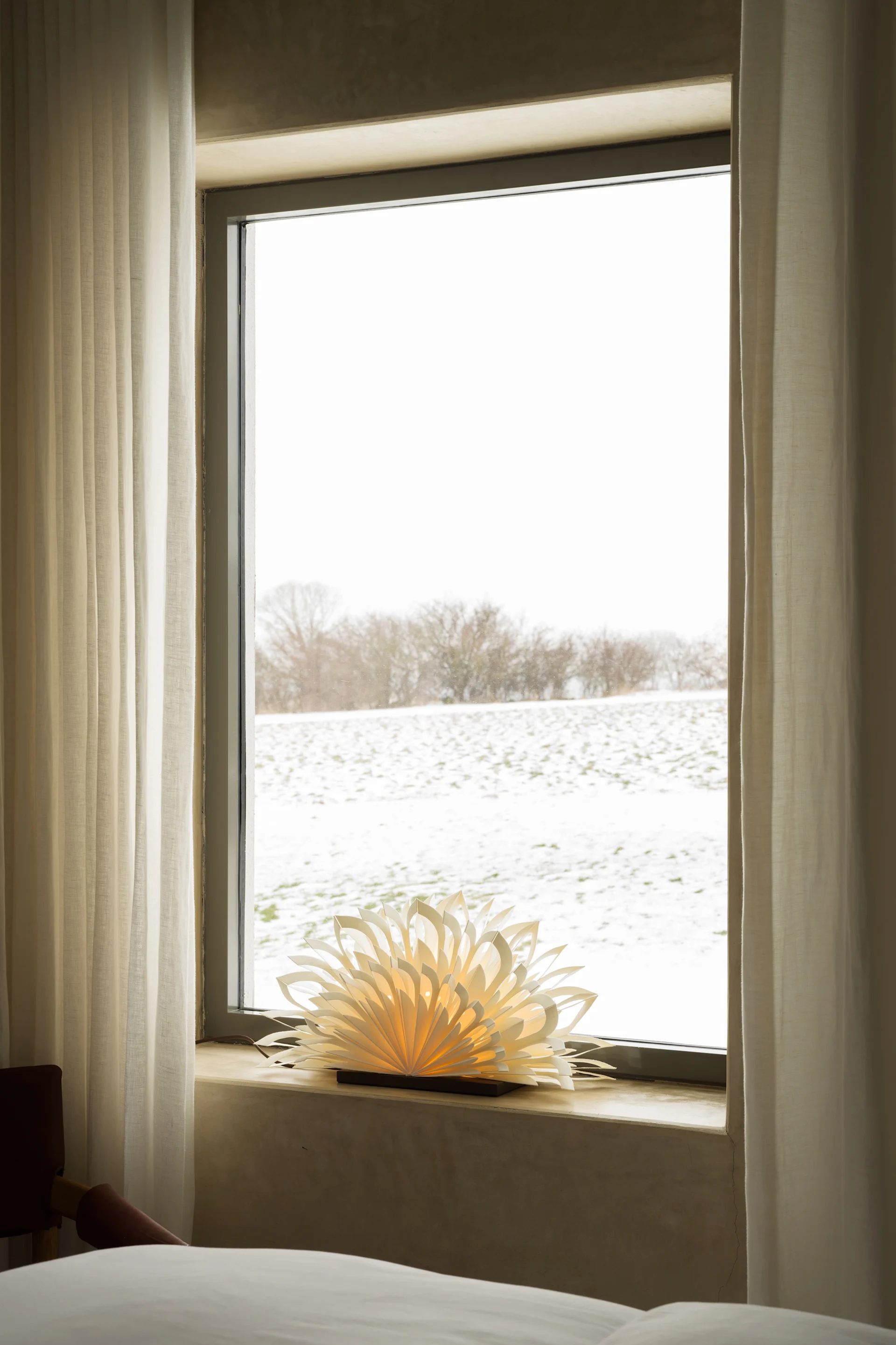 Vindu med utsikt over et snødekt felt, med hvite gardiner og en glødende papirlampe i vinduskarmen.