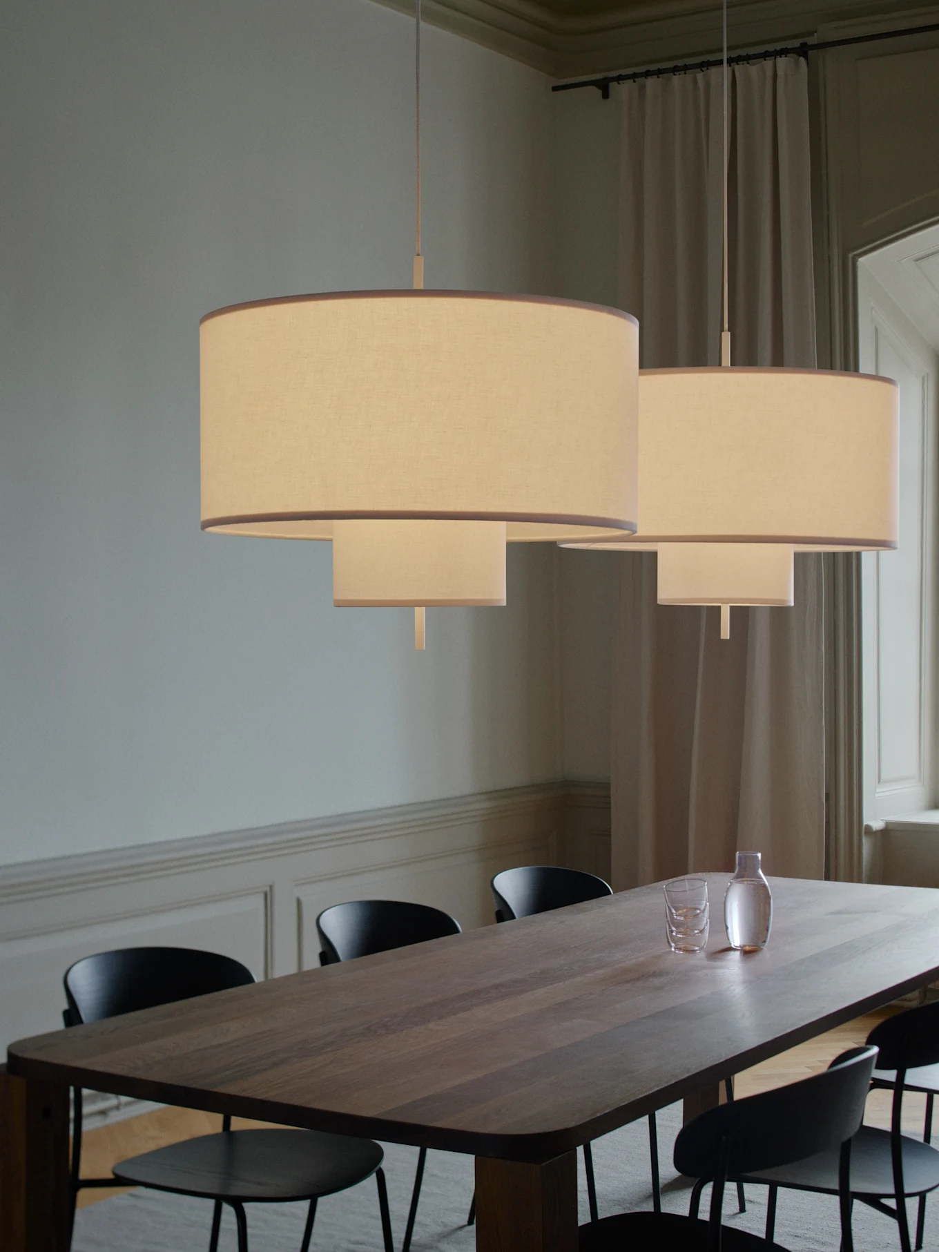 To etasjes stoffpendellamper lyser opp et langt spisebord i tre med svarte stoler, en karaffel og glass.