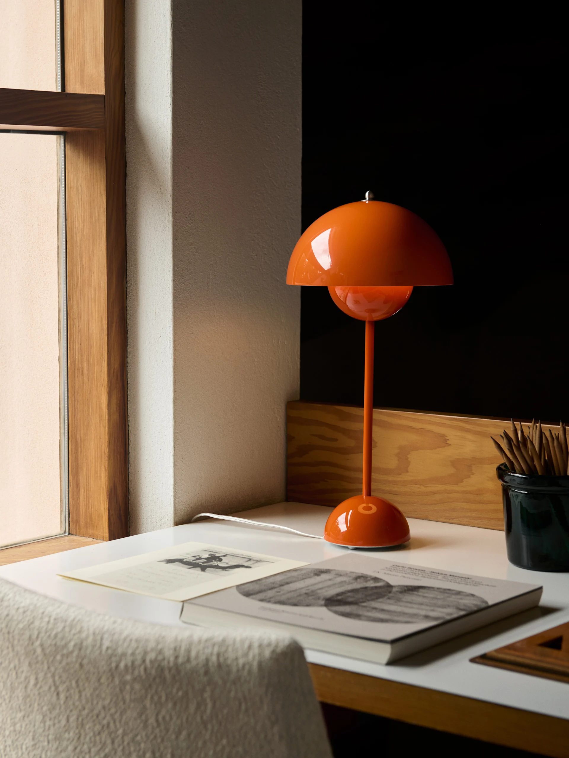 En oransje, kuppelformet bordlampe står på et hvitt skrivebord ved siden av bøker og en blyantbeholder.