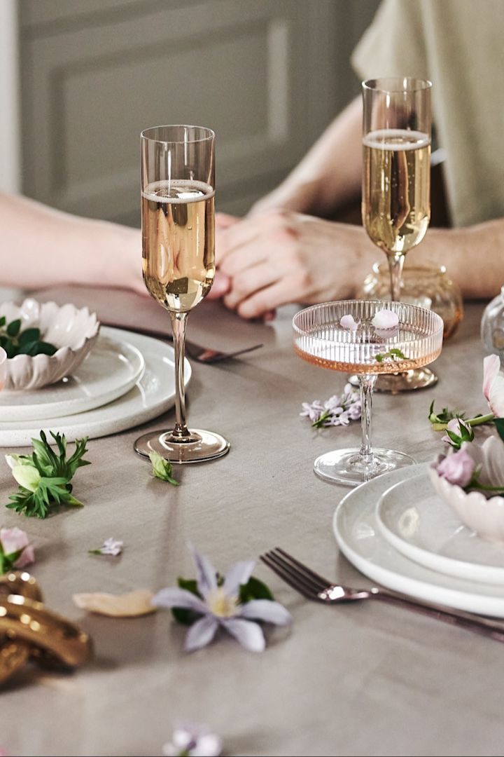 Til din romantiske middag hjemme kan du også dekke bordet med et lekent uttrykk. Her ser du hvitt servise, enkle glass og blomster på en beige duk.