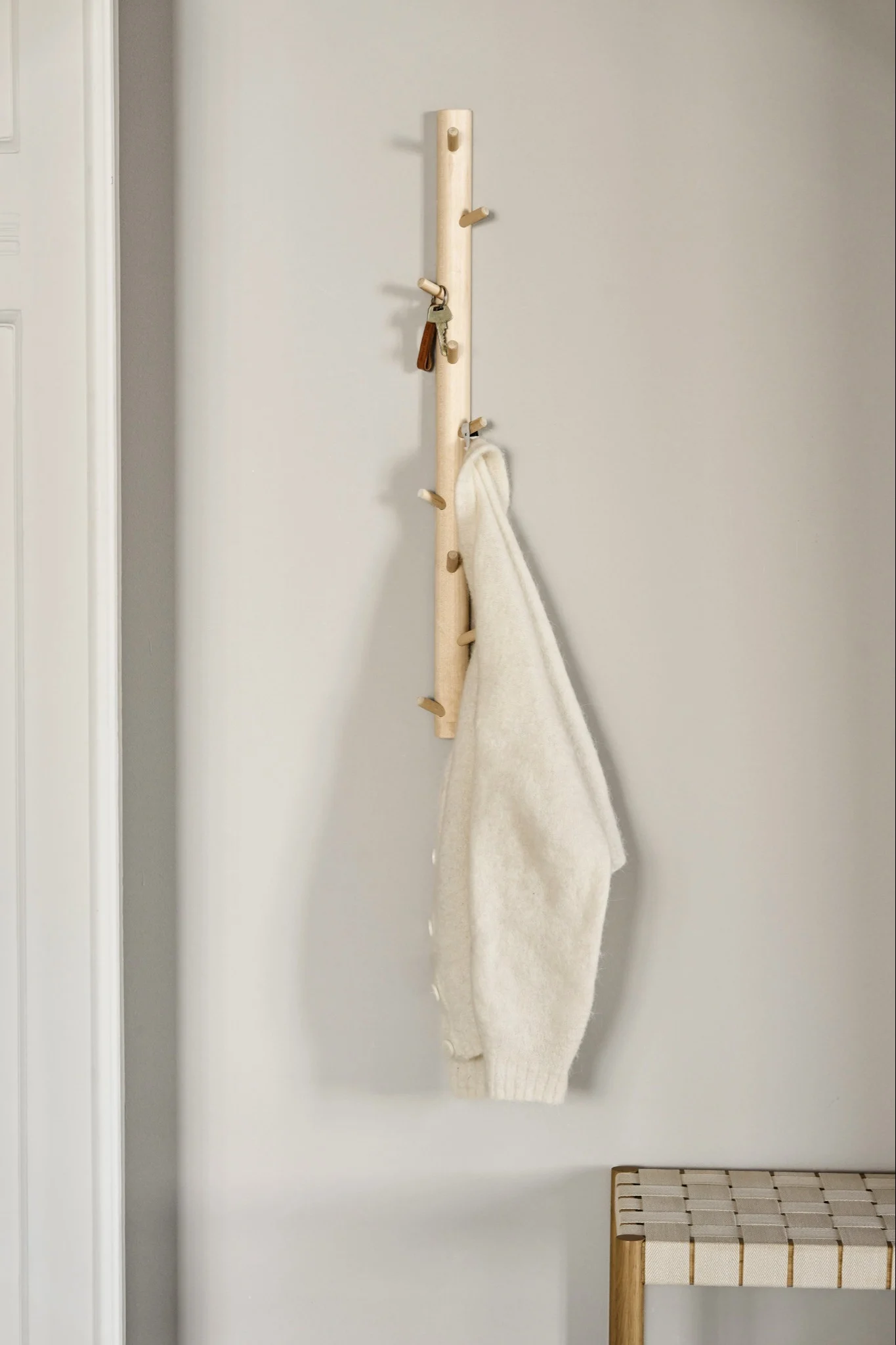 En lys kleshenger i tre med knagger holder nøkler og en kremfarget genser, med en flettet benk under.