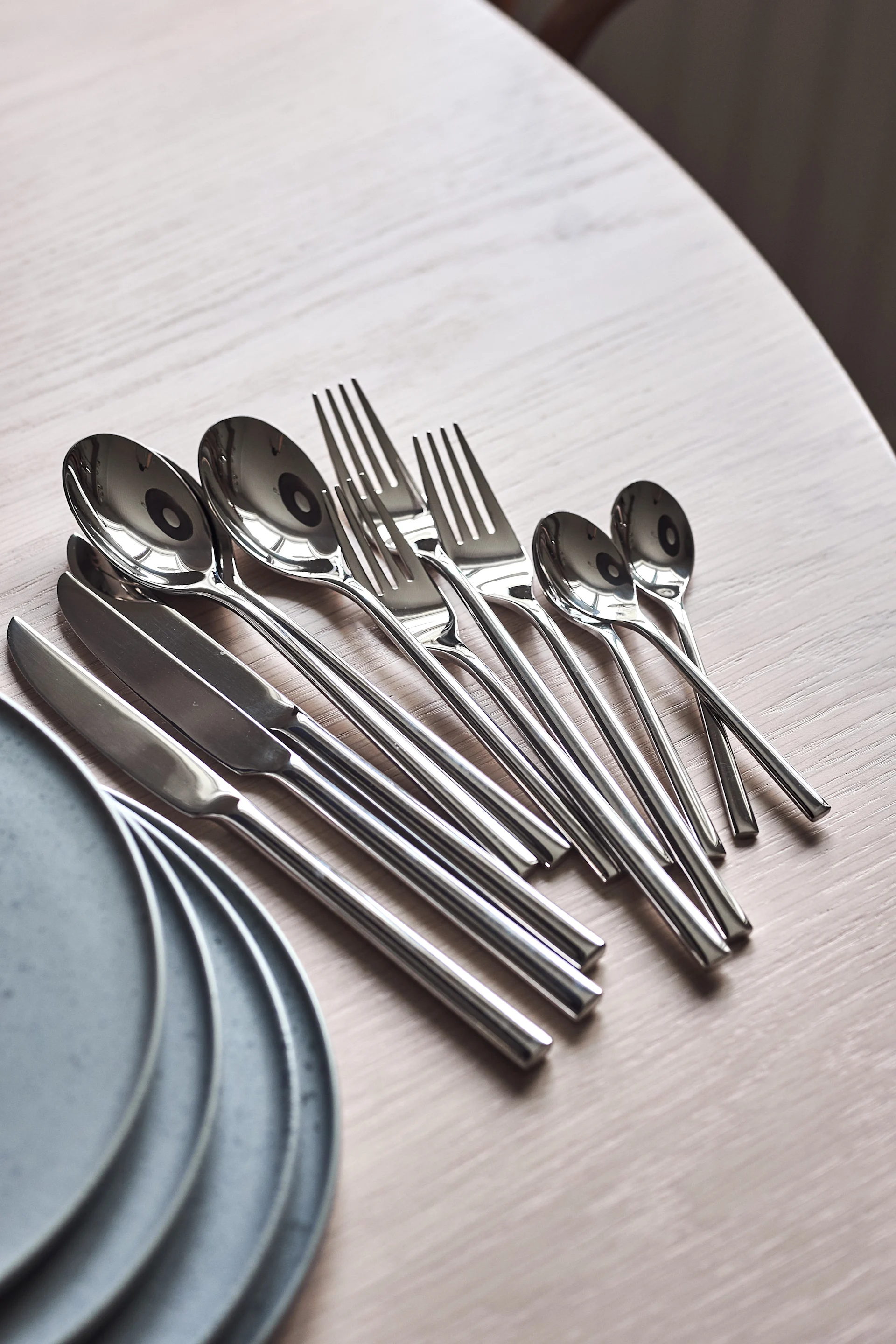 Sett med skinnende sølvfarget bestikk, inkludert skjeer, gafler og kniver, arrangert på et lyst trebord med grå tallerkener.
