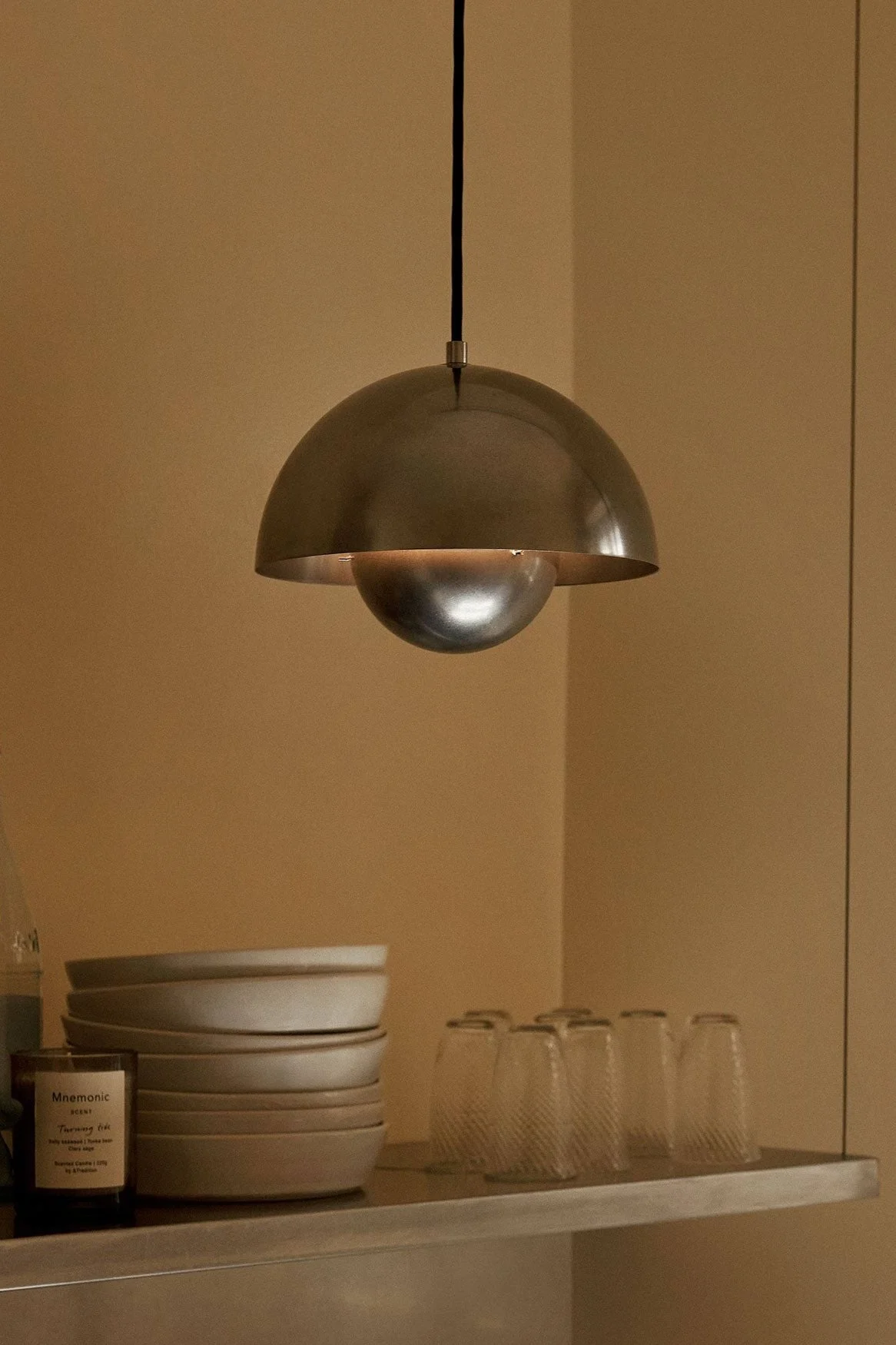 En kuppelformet metallpendellampe henger over en hylle med skåler, glass, flasker og et lys på en varm brun vegg.