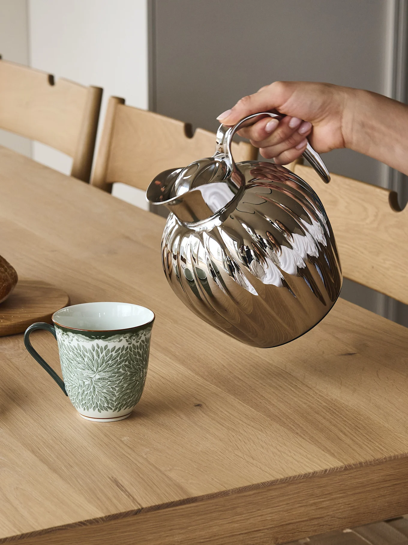 En hånd heller fra en sølvfarget riflet termokanne i et grønt og hvitt mønstret krus på et trebord.