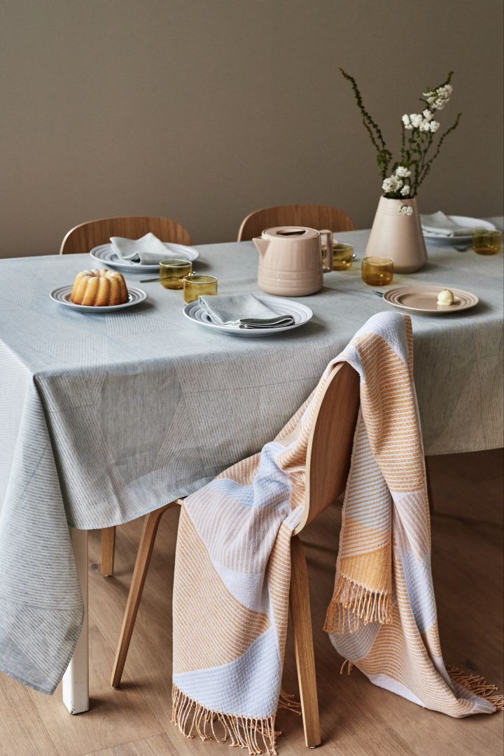 NJRD Stripes bomullspledd i blekgul og pastellblått drapert over en stol ved et spisebord. 