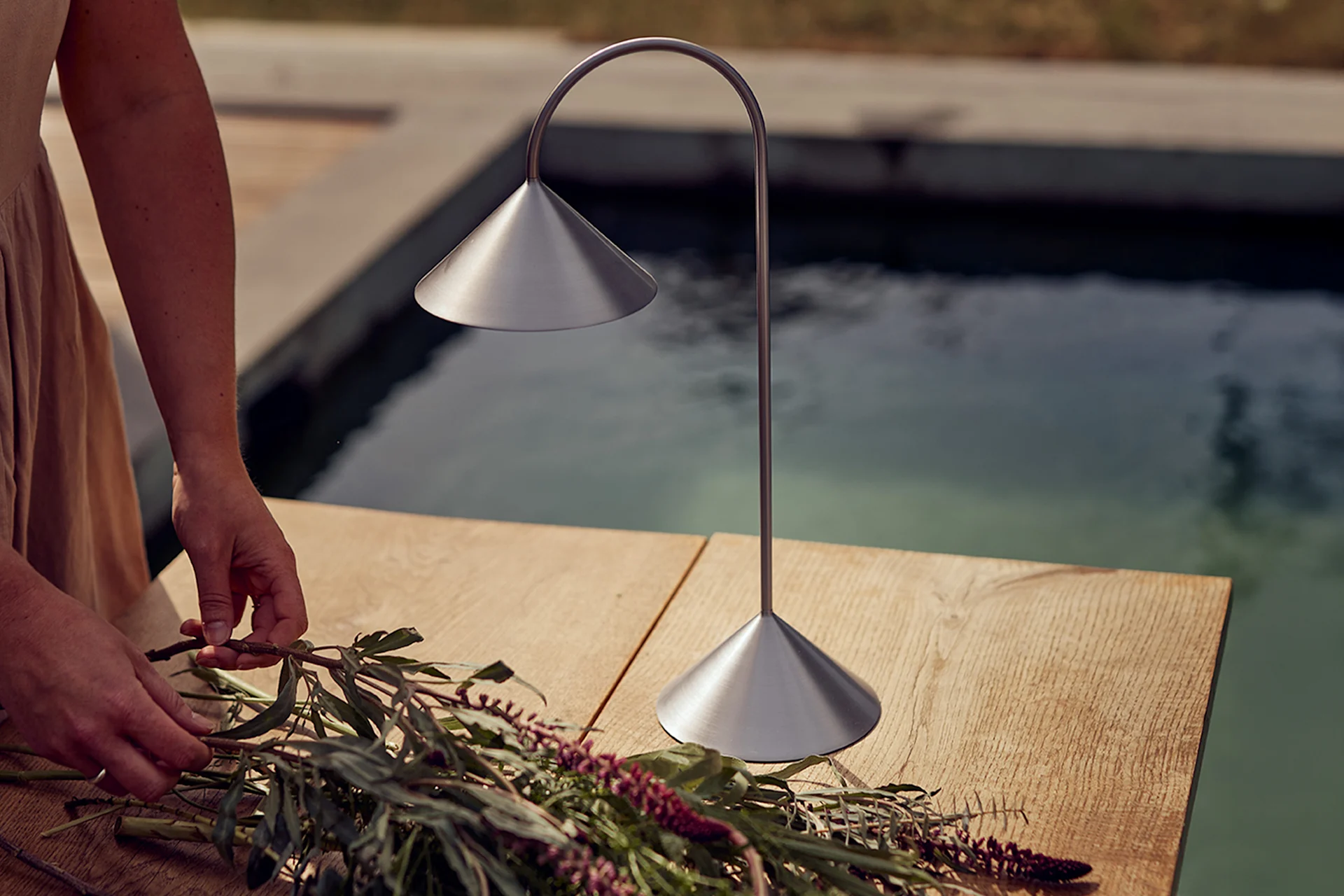 Hender arrangerer blomster på et trebord ved siden av en bordlampe i børstet metall ved et basseng.