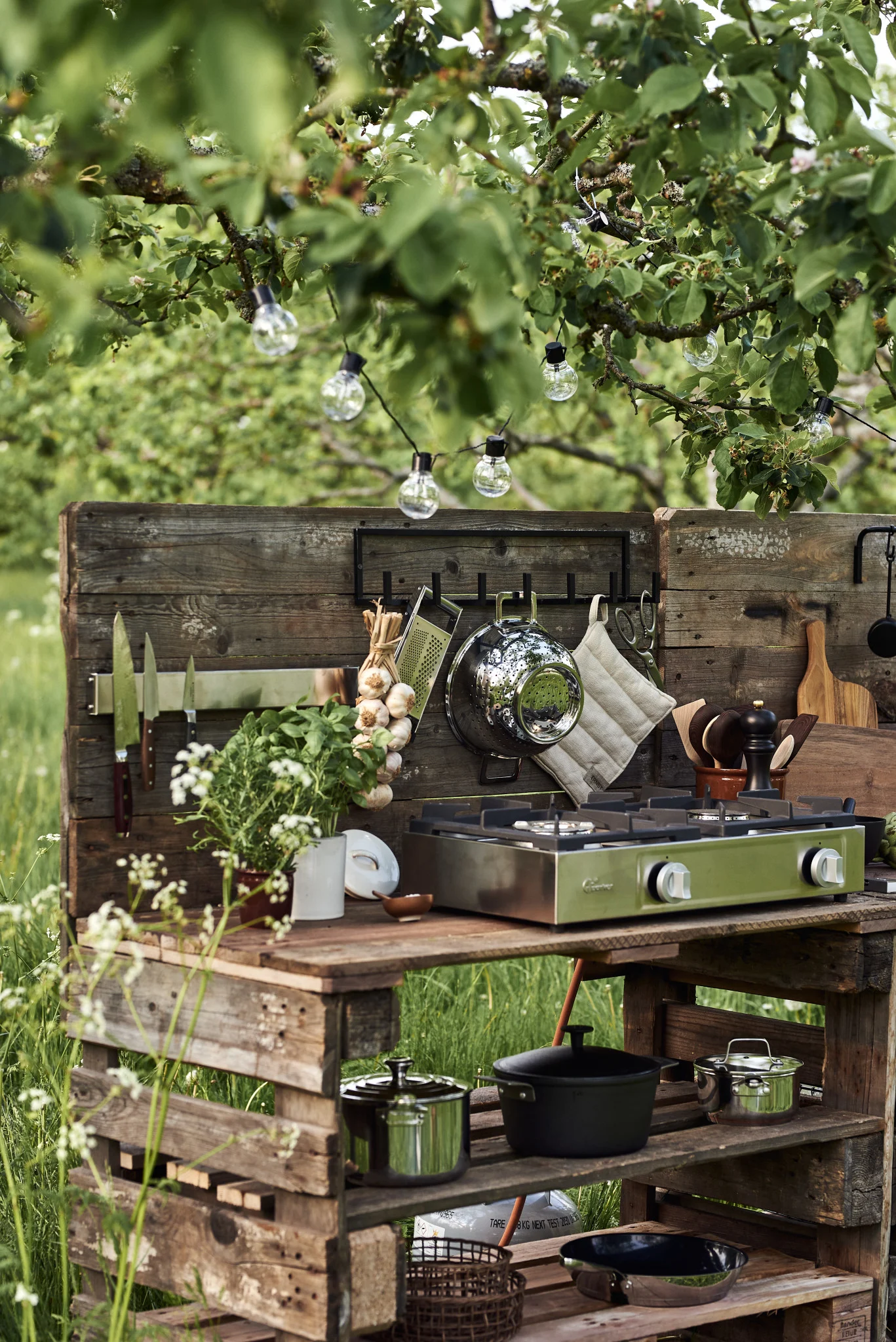 Et utekjøkken av trepaller med en grønn gasskomfyr med to brennere, hengende redskaper og potteurter under lyslenker.