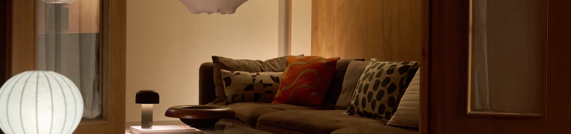 En koselig stue med en brun sofa, diverse mønstrede puter og to lamper: en stor hvit ribbet gulvlampe og en liten soppformet bordlampe.