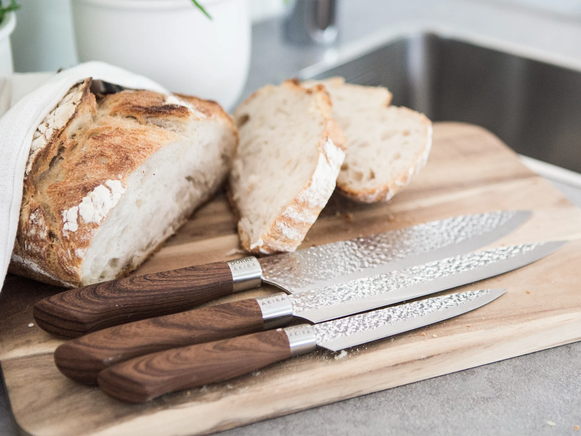 Nybakt rustikt brød, delvis skåret, på et skjærebrett av tre med tre kniver. Kjøkkenvask i bakgrunnen.