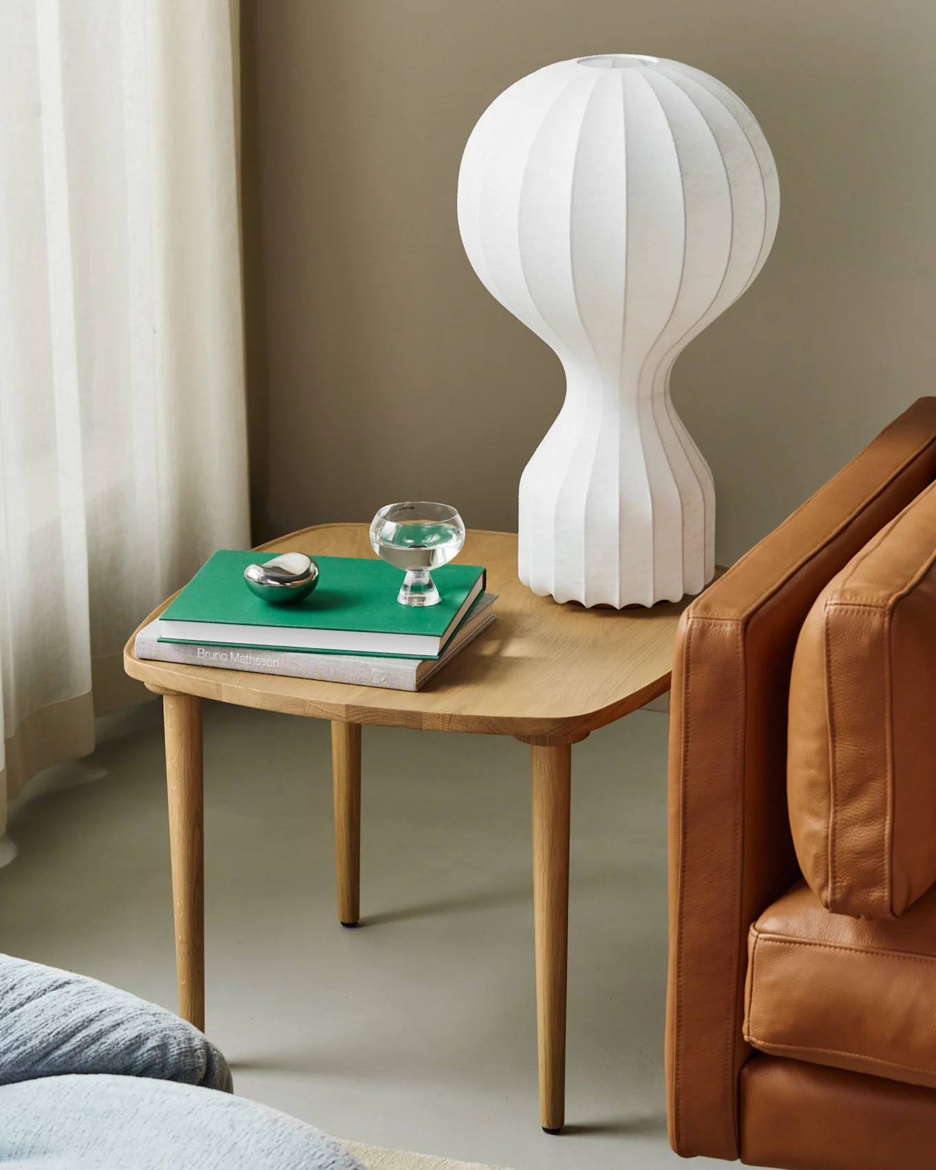 Tre sidebord med hvit riflet lampe, grønn bok, vannglass, sølvdekor ved siden av brun skinnsofa.